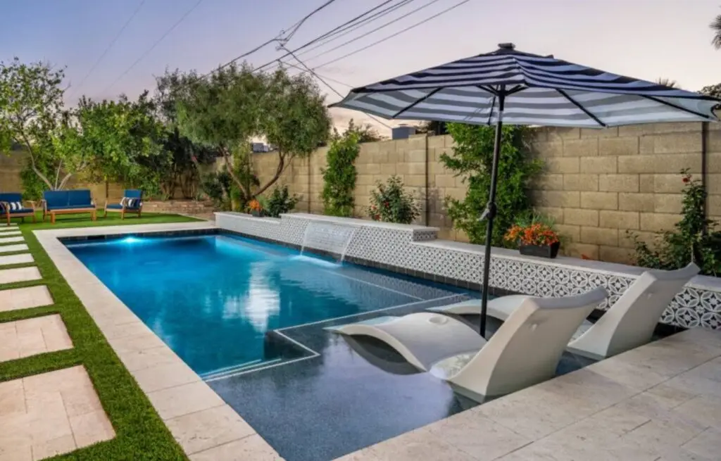 Modern backyard pool with water feature and built-in sun loungers under striped umbrella.